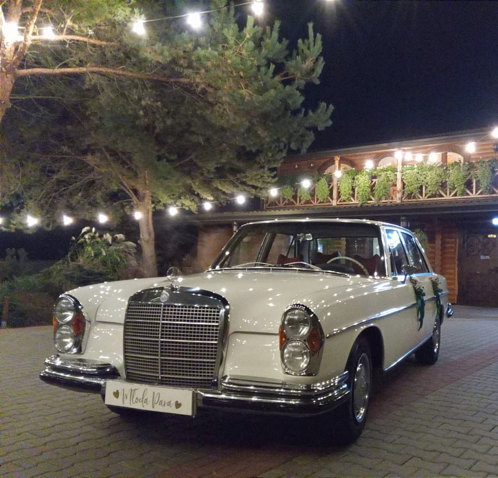 Obraz przedstawiający udekorowanego Mercedesa W108 stojącego przy restauracji, który prezentuje się w blasku oświetlenia parkingu
